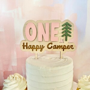 K&ouml;nnte beinhalten: Ein Holz-Kuchentopper mit dem Wort "ONE" in rosa Buchstaben und den Worten "Happy Camper" in braunen Buchstaben. Rechts neben dem Wort "ONE" befindet sich ein gr&uuml;ner Tannenbaum.