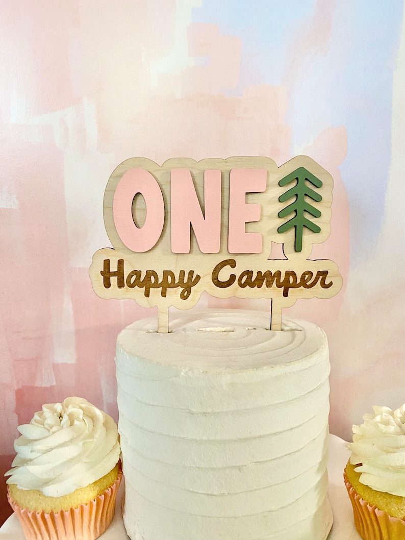 K&ouml;nnte beinhalten: Ein Holz-Kuchentopper mit dem Wort "ONE" in rosa Buchstaben und den Worten "Happy Camper" in braunen Buchstaben. Rechts neben dem Wort "ONE" befindet sich ein gr&uuml;ner Tannenbaum.