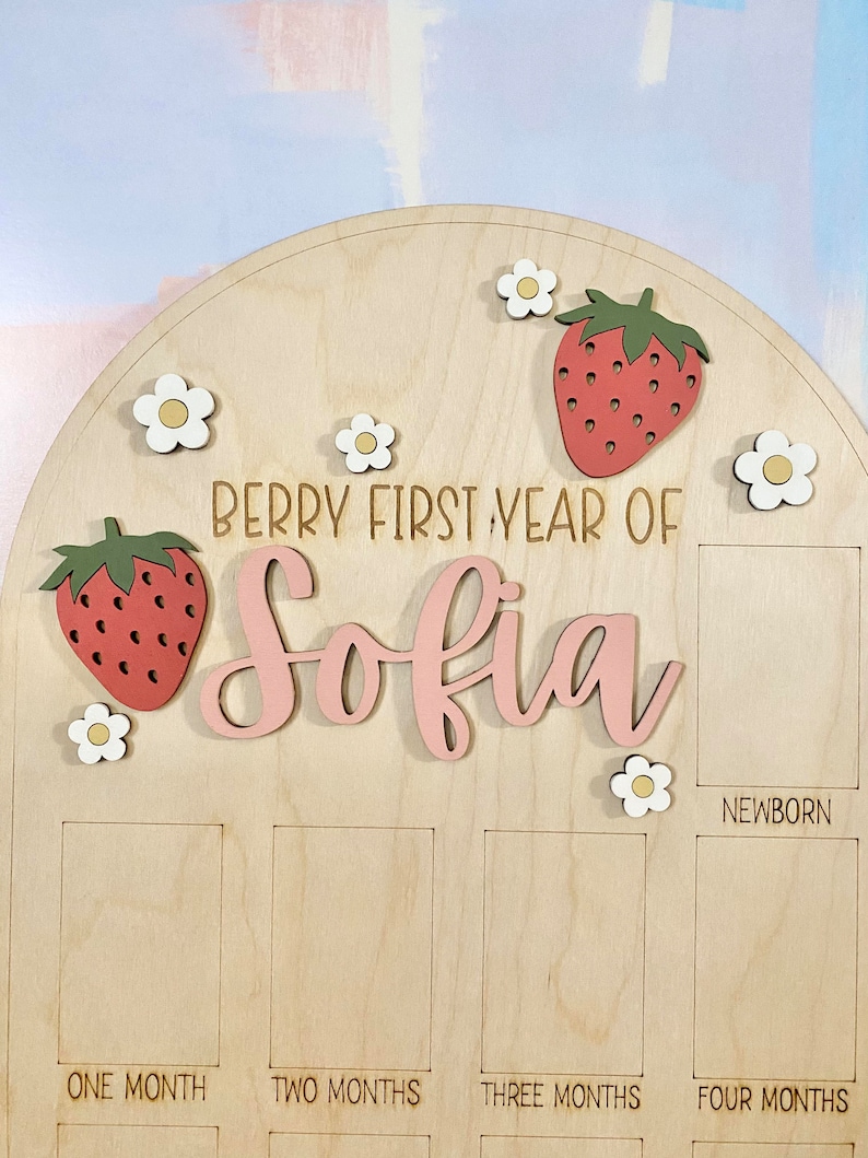 Puede incluir: Tablero de hitos de madera con el texto "Berry First Year of Sofia". Cuenta con recortes de fresas y flores. Incluye espacios para fotos etiquetados como "Newborn", "One Month", "Two Months", "Three Months" y "Four Months".