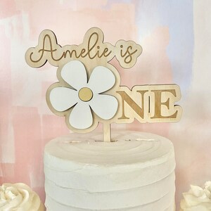 Pu&ograve; includere: Un cake topper in legno con la scritta "Amelie is ONE". Il topper presenta un design a margherita a strati con petali bianchi e un centro dorato. La torta &egrave; bianca con una superficie strutturata e ci sono cupcake visibili su entrambi i lati.