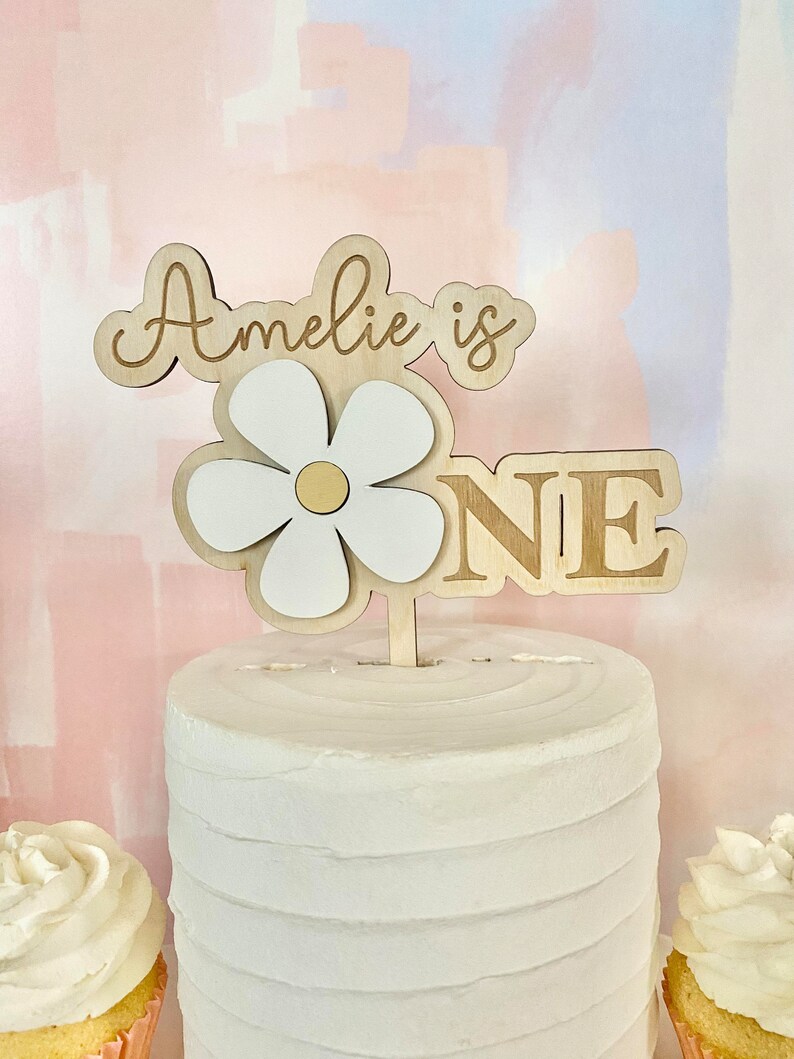 Pu&ograve; includere: Un cake topper in legno con la scritta "Amelie is ONE". Il topper presenta un design a margherita a strati con petali bianchi e un centro dorato. La torta &egrave; bianca con una superficie strutturata e ci sono cupcake visibili su entrambi i lati.