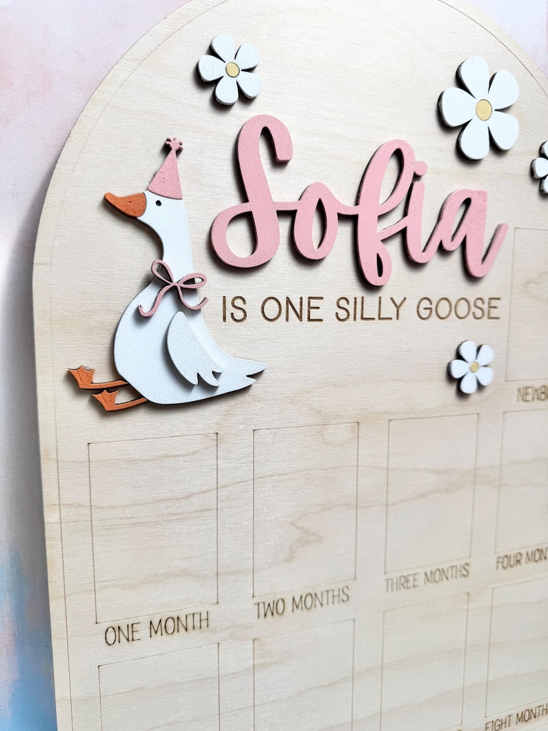 May include: Wooden baby milestone board with the name "Sofia" in pink script. A whimsical goose wearing a party hat and flowers decorate the board. The text "IS ONE SILLY GOOSE" is below the name. Monthly milestone boxes are etched into the wood.