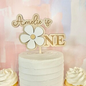 Pu&ograve; includere: Un cake topper in legno con le parole "Amelie is ONE" e un design a margherita. Il cake topper &egrave; posto su una torta glassata bianca, con due cupcake su entrambi i lati.