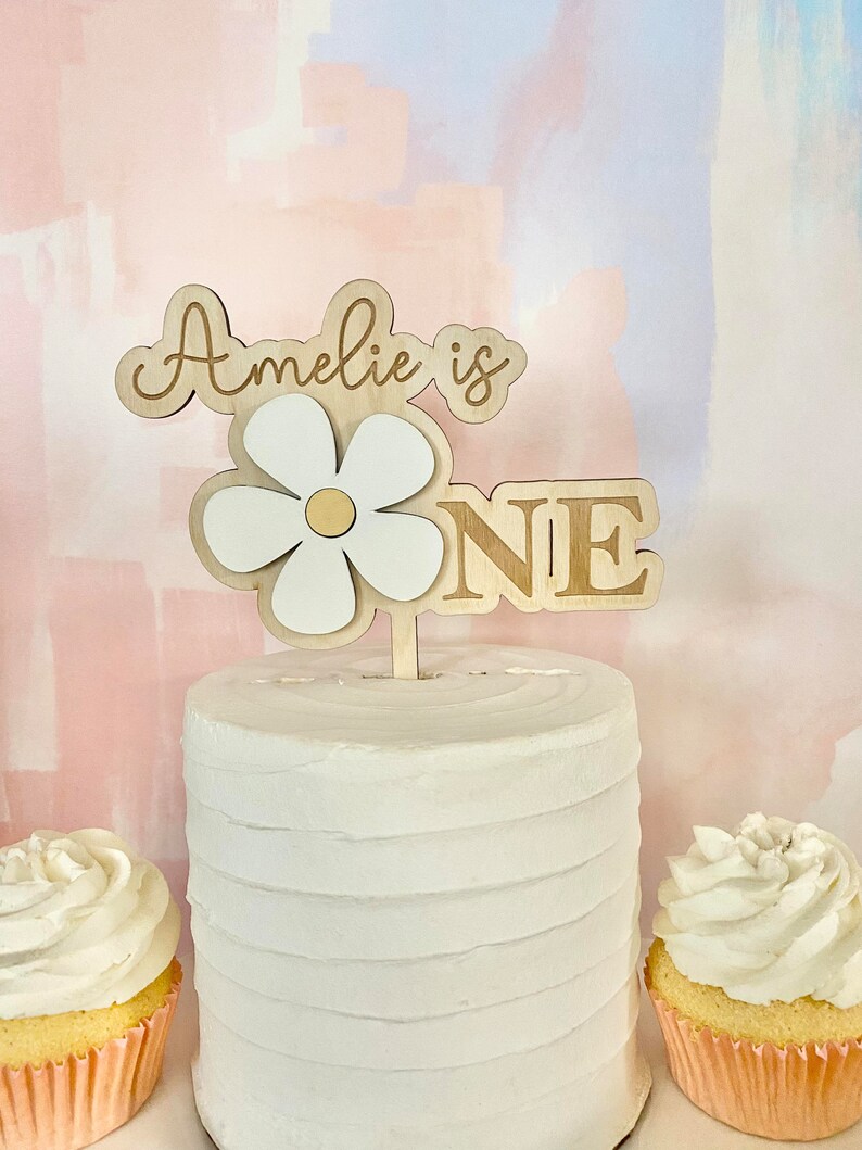 Pu&ograve; includere: Un cake topper in legno con le parole "Amelie is ONE" e un design a margherita. Il cake topper &egrave; posto su una torta glassata bianca, con due cupcake su entrambi i lati.