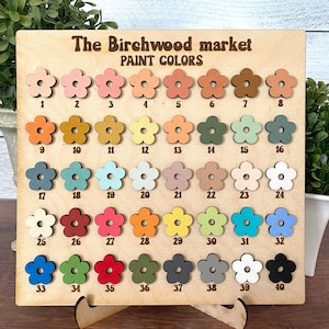 Pu&ograve; includere: Tabella colori in legno di The Birchwood Market. La tabella presenta 40 campioni di vernice a forma di fiore in vari colori, numerati da 1 a 40. Il testo "PAINT COLORS" &egrave; visualizzato sopra i campioni.