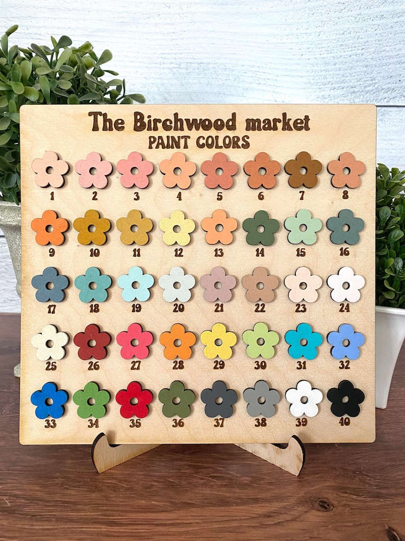 Pu&ograve; includere: Tabella colori in legno di The Birchwood Market. La tabella presenta 40 campioni di vernice a forma di fiore in vari colori, numerati da 1 a 40. Il testo "PAINT COLORS" &egrave; visualizzato sopra i campioni.