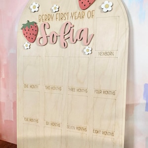 Puede incluir: Tablero de hitos de madera clara con una parte superior arqueada. El texto "Berry First Year of Sofia" es rosa, con fresas y flores decorativas. Tiene espacios para fotos mensuales para rastrear el primer a&ntilde;o del beb&eacute;.