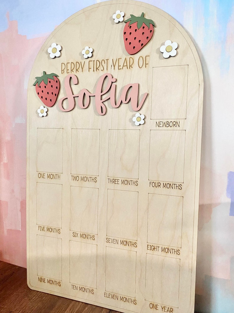 Puede incluir: Tablero de hitos de madera clara con una parte superior arqueada. El texto "Berry First Year of Sofia" es rosa, con fresas y flores decorativas. Tiene espacios para fotos mensuales para rastrear el primer a&ntilde;o del beb&eacute;.