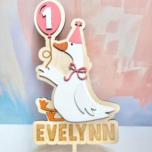 May include: Wooden cake topper featuring a white goose wearing a party hat and holding a pink balloon with the number 1. The goose has orange feet and a pink bow. The name "EVELYNN" is displayed on a wooden sign below.