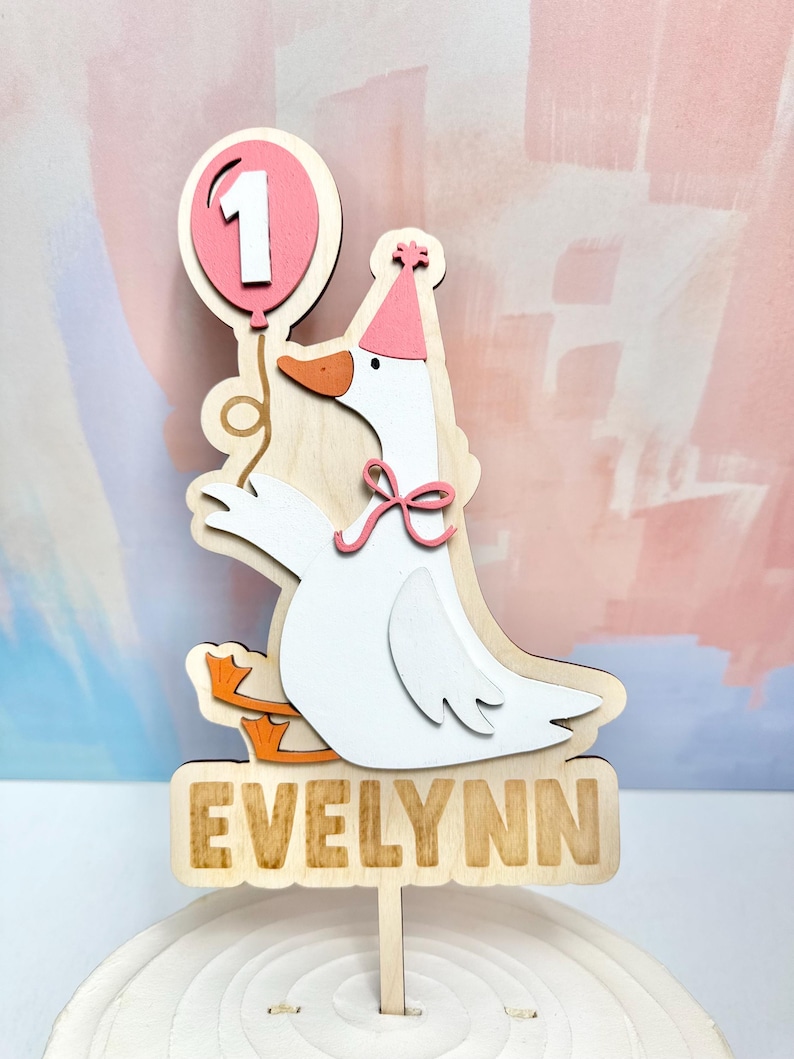 May include: Wooden cake topper featuring a white goose wearing a party hat and holding a pink balloon with the number 1. The goose has orange feet and a pink bow. The name "EVELYNN" is displayed on a wooden sign below.