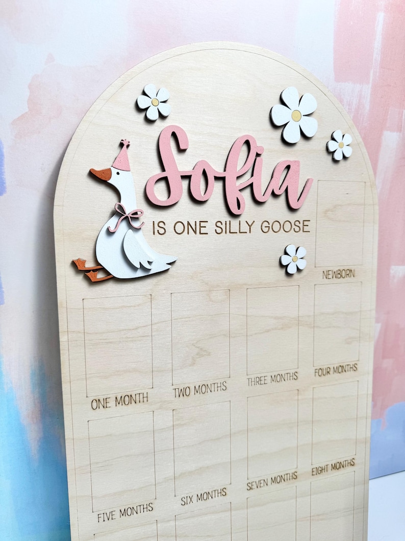 May include: Wooden milestone board with the name "Sofia" in pink script, a cartoon goose, and flower accents. The board is designed to track a baby's first year, with labeled boxes for each month.