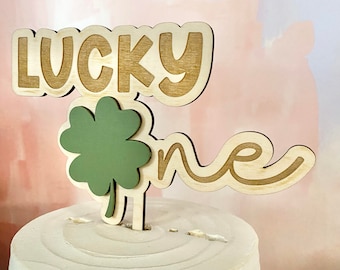 May include: A wooden cake topper with the words "Lucky One" in brown script. A green four-leaf clover is attached to the topper.