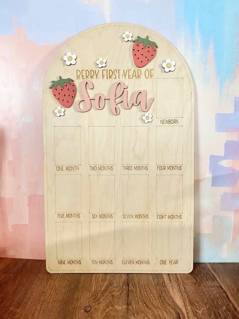 Puede incluir: Tablero de hitos de madera con tema de fresas y margaritas. El tablero tiene forma de arco en la parte superior y presenta el texto "Berry First Year of Sofia". Tiene espacios para fotos desde reci&eacute;n nacido hasta un a&ntilde;o.