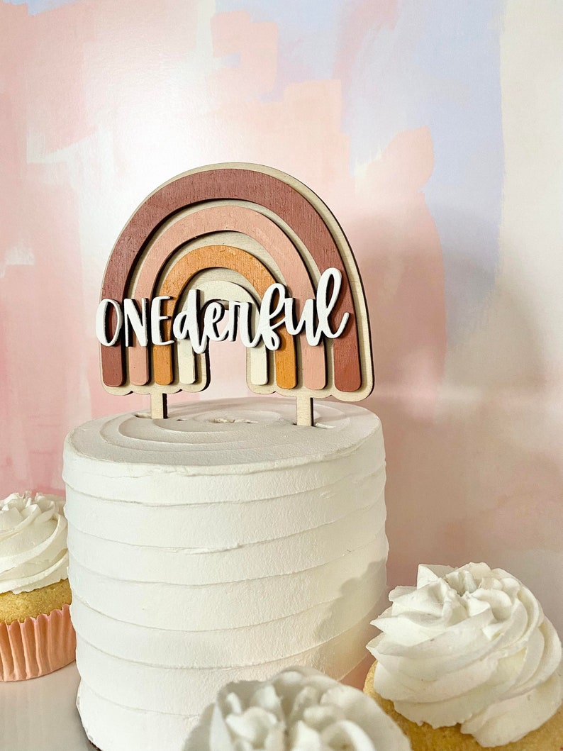 May include: A wooden rainbow cake topper with the word "ONEDERFUL" in white lettering. The rainbow is made of layered wood in shades of brown, pink, and orange.