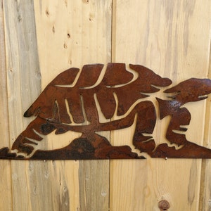 May include: A rusty metal turtle silhouette wall decor. The turtle is facing right and has a detailed shell and legs. The decor is mounted on a light wood background.