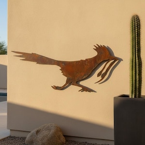 May include: A rust-colored metal wall art piece shaped like a roadrunner bird in a running pose. The silhouette features a long tail, a crest on its head, and a slender beak. The metal has a weathered, oxidized finish with varied brown tones.