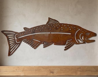 May include: A flat, rust-colored metal wall art piece shaped like a trout. The fish silhouette features cut-out details along the body, fins, and tail, with a textured, weathered surface finish that mimics the appearance of oxidized iron metal.