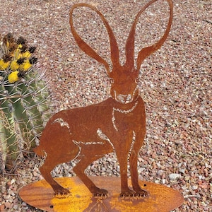May include: A rusty metal sculpture of a jackrabbit with long ears. The sculpture is standing on a flat, rusty metal base. The sculpture is in front of a cactus.