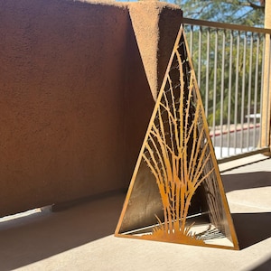 May include: A triangular metal sculpture with a cutout design of a tree. The sculpture is made of a gold-colored metal and has a rustic, weathered look.