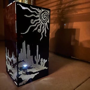 May include: A black metal lantern with a cut-out design of a desert scene, including a sun, cacti, and mountains. The lantern is lit from within, casting a warm glow.