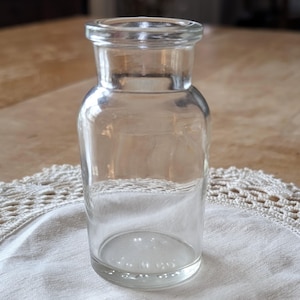 Puede incluir: Un tarro de farmacia de vidrio transparente con un cuerpo cilíndrico y un cuello corto. El tarro está vacío y se encuentra sobre un tapete blanco con bordes de encaje. El fondo es una superficie marrón claro.