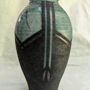 Puede incluir: Un jarrón de cerámica hecho a mano con una combinación de colores verde azulado oscuro y negro. El jarrón presenta una superficie texturizada y un diseño de líneas negras verticales y formas geométricas. El jarrón mide unos 25 cm de altura.