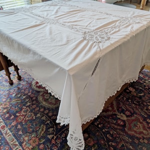 May include: A white rectangular tablecloth with intricate lace detailing. The tablecloth is draped over a wooden table, with the lace trim visible along the edges. The design includes a central pattern and corner accents.