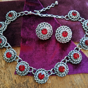 Könnte beinhalten: Eine silberne Halskette mit roten Strasssteinen und passenden Ohrringen. Die Halskette hat ein gedrehtes Kettenmuster und die Ohrringe sind rund mit einem gedrehten Metalldesign.