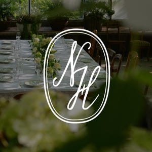 Könnte beinhalten: Ein langer Esstisch mit weißen Tellern, klaren Gläsern und Blumenarrangements, gesehen durch einen unscharfen Vordergrund aus weißen Blüten. Ein weißes ovales Logo mit den stilisierten, ineinander verschlungenen Buchstaben SH ist zentriert.