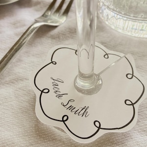 Puede incluir: Una tarjeta de mesa blanca con un borde decorativo negro y el nombre "Jacob Smith" en escritura cursiva. Una copa de vino transparente se encuentra sobre la tarjeta, con un tenedor plateado y un vaso de agua en el fondo.