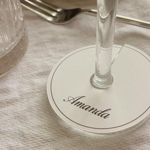 Könnte beinhalten: Ein runder Weinglas-Anhänger aus weißem Acryl mit einem dünnen schwarzen Rand und dem Namen Amanda in schwarzer Schreibschrift. Der Anhänger hat einen kleinen Schlitz, um ihn am Stiel eines Glases zu befestigen und das Getränk zu kennzeichnen.