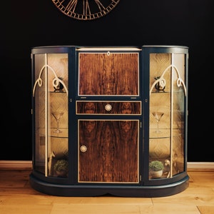May include: A dark blue, curved cabinet with glass doors and a wooden centre panel. The cabinet has gold trim and decorative details. The cabinet is sitting on a wooden floor.