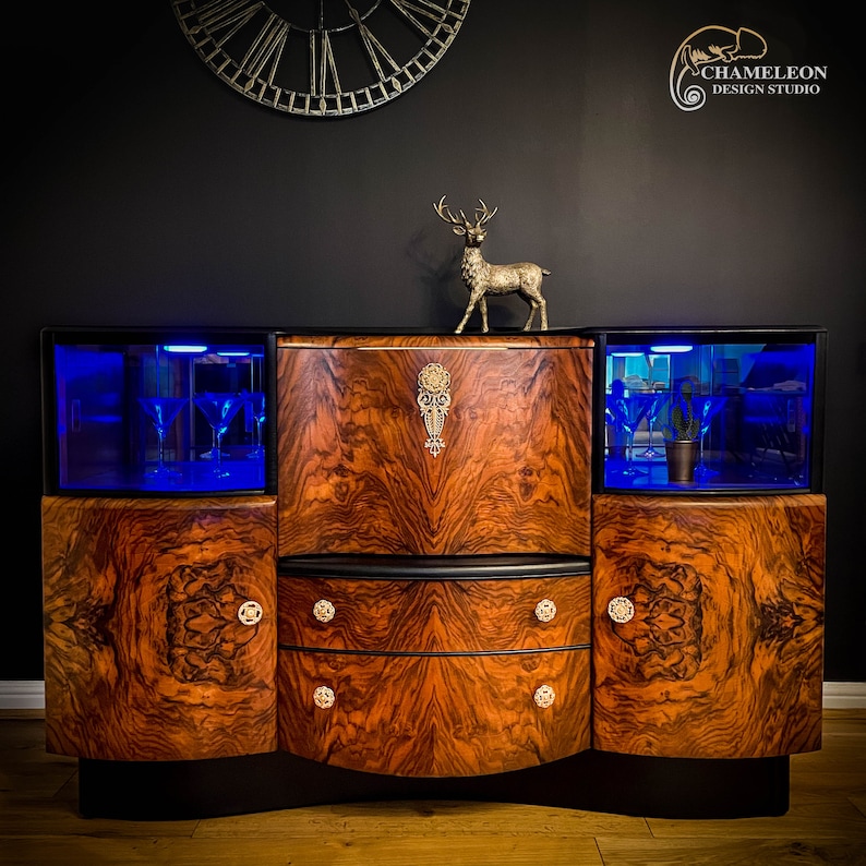 May include: A vintage wooden bar cabinet with a black finish and two glass doors. The cabinet has a curved front and two drawers with ornate metal handles. The cabinet is lit with blue lights and features a decorative metal design on the top.