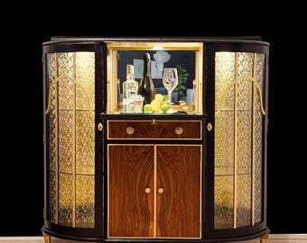 May include: A black wooden bar cabinet with glass doors and a mirrored back. The cabinet has a single drawer and two doors with gold accents. The interior is lit and contains bottles of liquor and glasses.