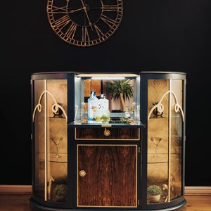 May include: A dark blue, curved cabinet with glass doors and gold accents. The cabinet has a wooden interior with a shelf and a drawer. There are two bottles of spirits and a glass on the shelf. The cabinet is in front of a black wall with a gold clock.