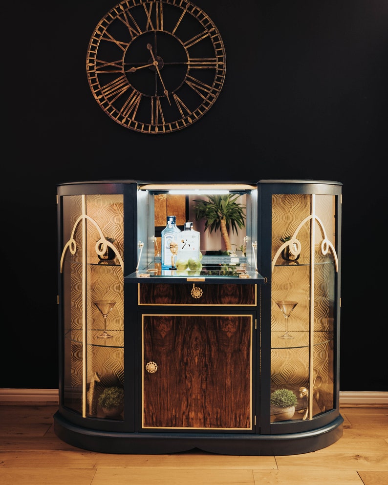 May include: A dark blue, curved cabinet with glass doors and gold accents. The cabinet has a wooden interior with a shelf and a drawer. There are two bottles of spirits and a glass on the shelf. The cabinet is in front of a black wall with a gold clock.
