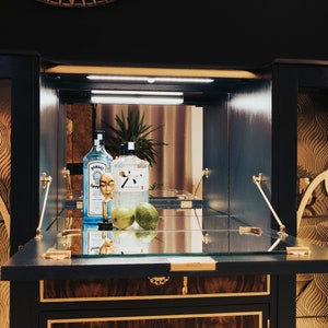 May include: A dark blue wooden bar cabinet with a glass top and gold accents. The cabinet is open, revealing two clear glass bottles, a gold bottle stopper, and two green limes. The bottles are labelled "Bombay Sapphire" and "Tanqueray Gin".