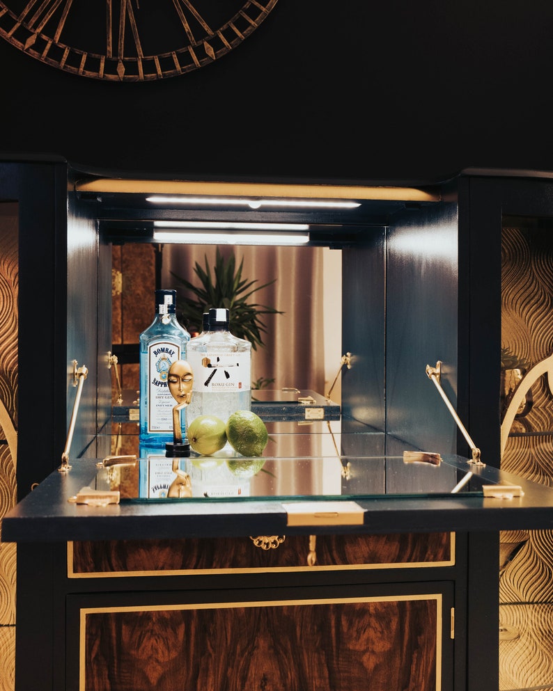 May include: A dark blue wooden bar cabinet with a glass top and gold accents. The cabinet is open, revealing two clear glass bottles, a gold bottle stopper, and two green limes. The bottles are labelled "Bombay Sapphire" and "Tanqueray Gin".