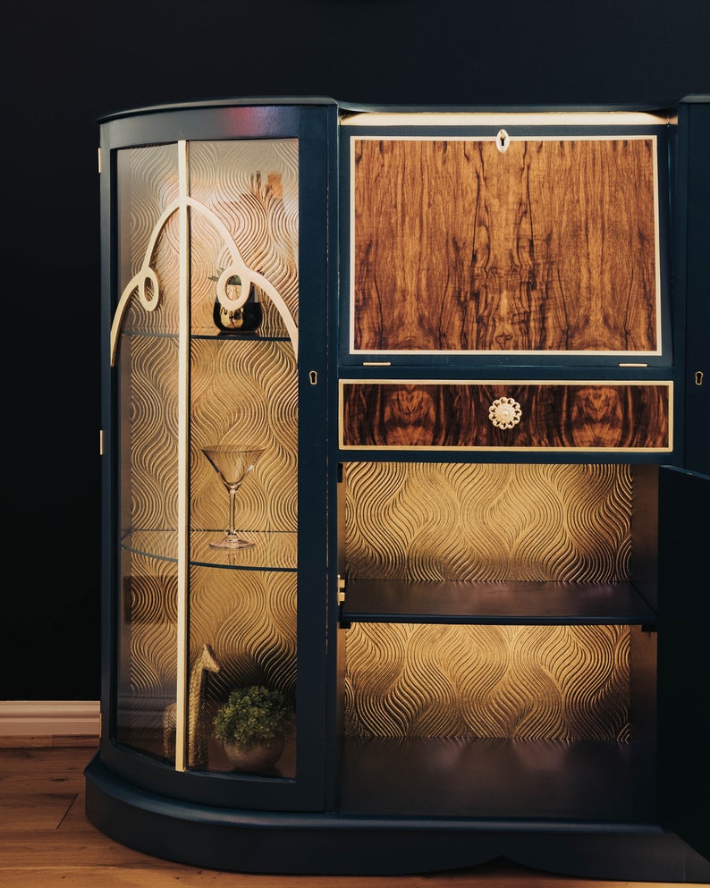 May include: A dark blue wooden cabinet with glass doors and shelves. The cabinet has a gold trim and a patterned wallpaper lining the shelves. The cabinet has a closed wooden door on the right side.