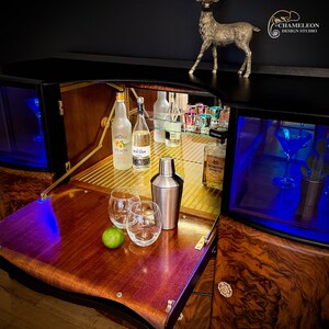 May include: A wooden bar cabinet with a black finish and a mirrored interior. The cabinet has a pull-out tray with two glasses, a lime, and a cocktail shaker. The cabinet also has a glass shelf with bottles of liquor and a glass of liquor.