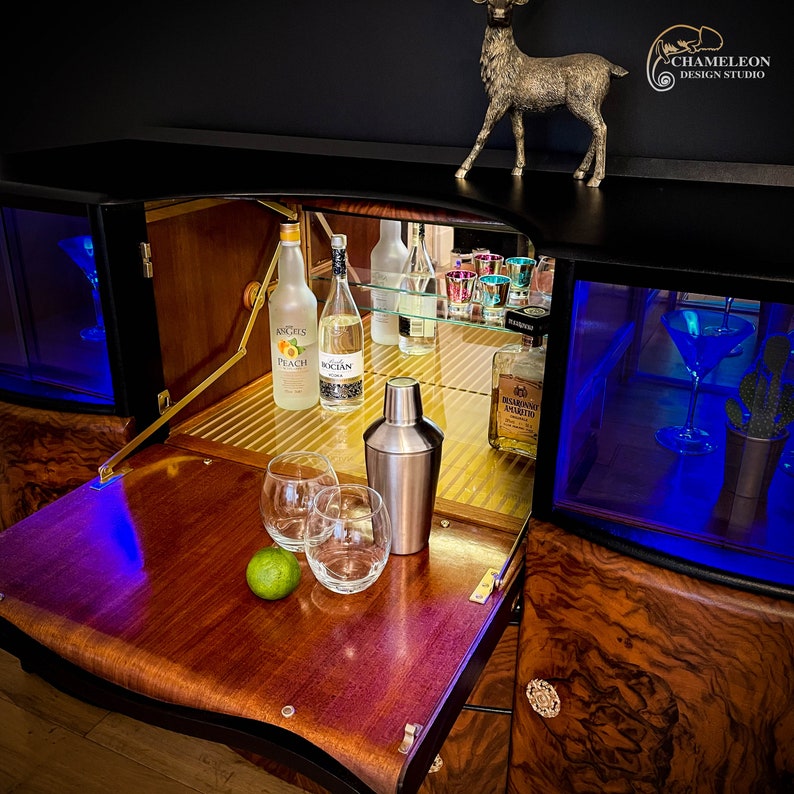 May include: A wooden bar cabinet with a black finish and a mirrored interior. The cabinet has a pull-out tray with two glasses, a lime, and a cocktail shaker. The cabinet also has a glass shelf with bottles of liquor and a glass of liquor.