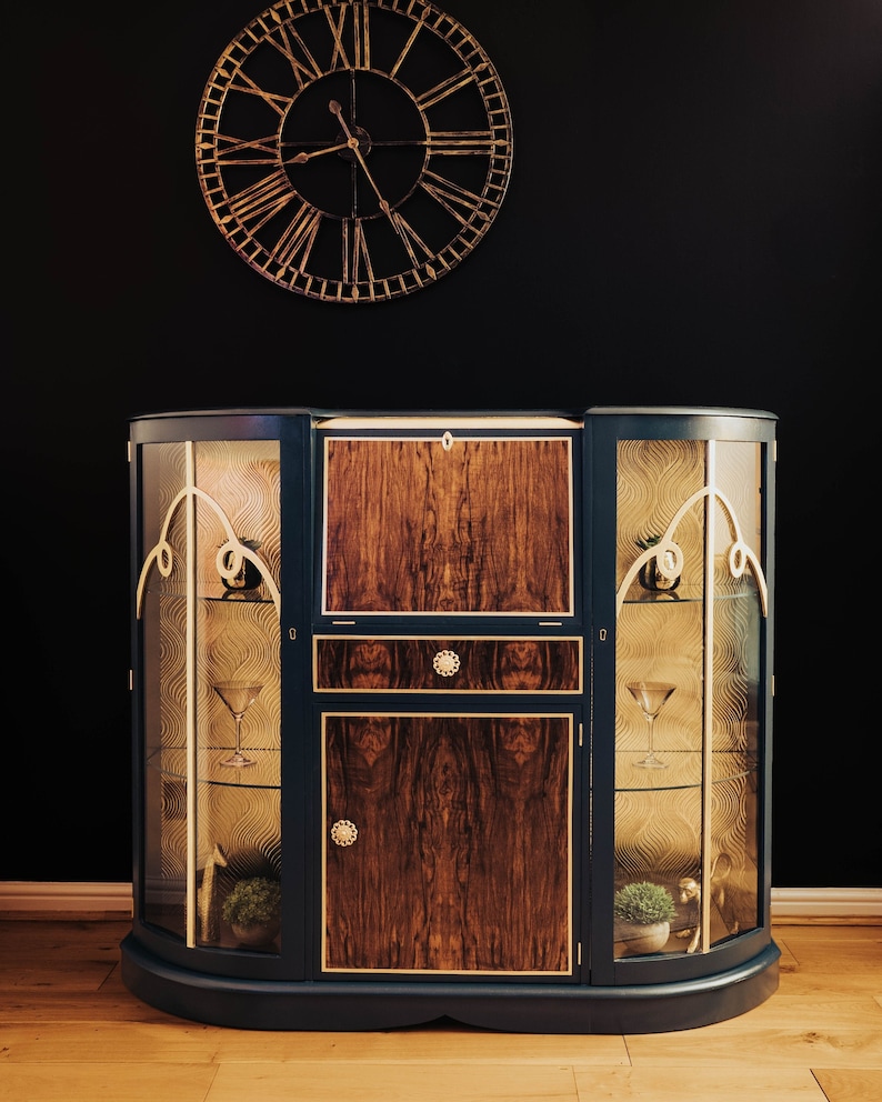 May include: A dark blue, curved cabinet with glass doors and a wooden centre panel. The cabinet has gold trim and decorative details. The cabinet is sitting on a wooden floor.