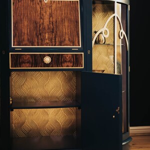 May include: A dark blue wooden cabinet with a glass door and a wood panel door. The cabinet has a gold-coloured trim and a gold-coloured design on the glass door. The cabinet has two shelves inside and a drawer on the top. The cabinet is open and the shelves are empty.