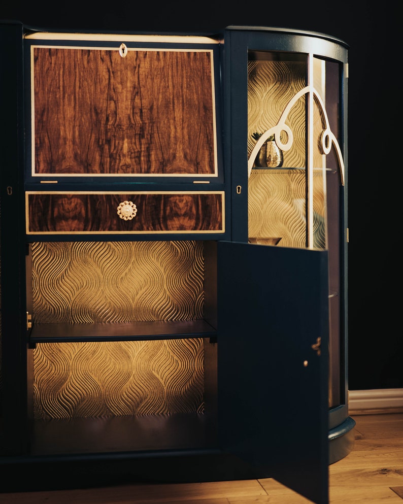 May include: A dark blue wooden cabinet with a glass door and a wood panel door. The cabinet has a gold-coloured trim and a gold-coloured design on the glass door. The cabinet has two shelves inside and a drawer on the top. The cabinet is open and the shelves are empty.