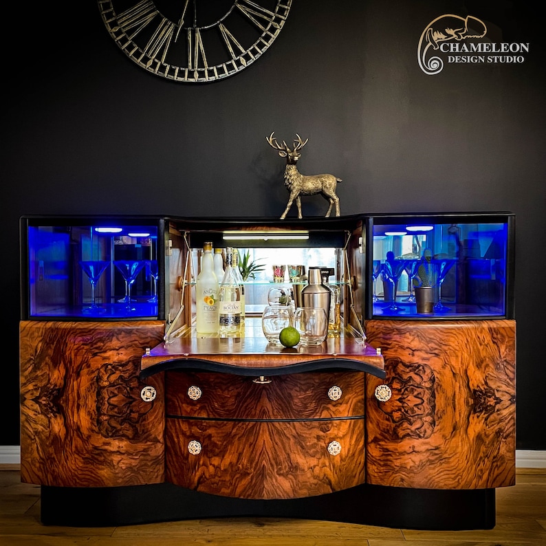 May include: A wooden bar cabinet with a black top and two glass doors on each side. The cabinet has a mirrored back and a shelf inside. The cabinet has a black base and two drawers. The cabinet is decorated with a wood grain pattern. The cabinet is lit with blue LED lights. The cabinet is sitting on a wooden floor. The text "CHAMELEON DESIGN STUDIO" is visible in the top right corner of the image.