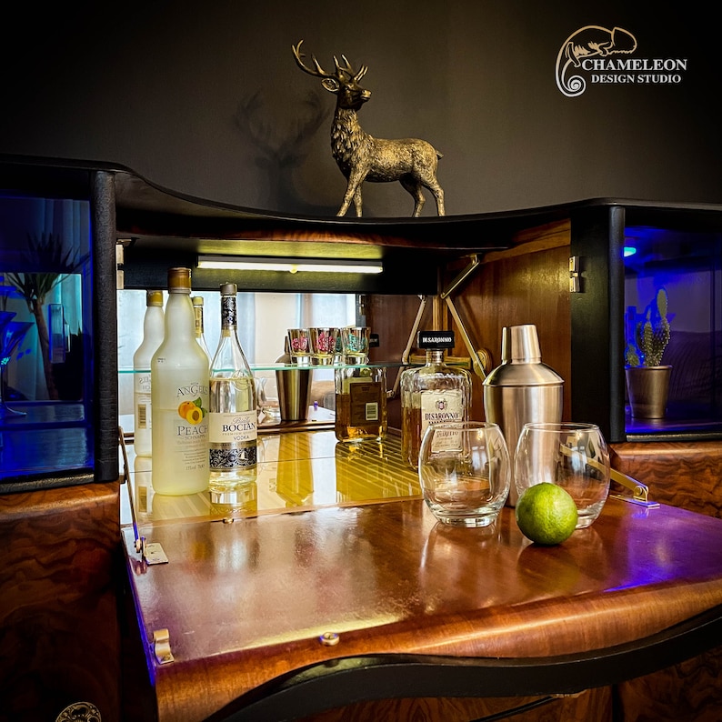 May include: A wooden bar cabinet with a black top and a mirrored interior. The cabinet has a fold-down bar top with a variety of bottles, glasses, and a cocktail shaker. The cabinet is decorated with a deer statue and the text "Chameleon Design Studio".