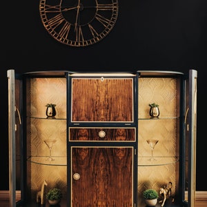 May include: A dark blue, curved cabinet with gold accents and glass shelves. The cabinet has two doors on either side and a centre section with a drawer and two doors. The cabinet is decorated with a gold, geometric pattern on the inside of the doors.
