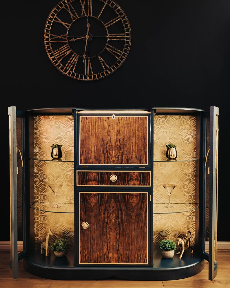 May include: A dark blue, curved cabinet with gold accents and glass shelves. The cabinet has two doors on either side and a centre section with a drawer and two doors. The cabinet is decorated with a gold, geometric pattern on the inside of the doors.
