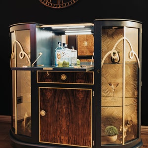 May include: A dark blue, curved, wooden bar cabinet with glass doors and shelves. The cabinet has gold accents and a wood-grain pattern. The interior of the cabinet is lit and contains bottles of spirits and limes.