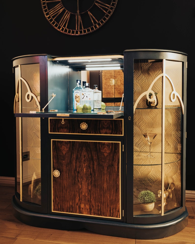 May include: A dark blue, curved, wooden bar cabinet with glass doors and shelves. The cabinet has gold accents and a wood-grain pattern. The interior of the cabinet is lit and contains bottles of spirits and limes.